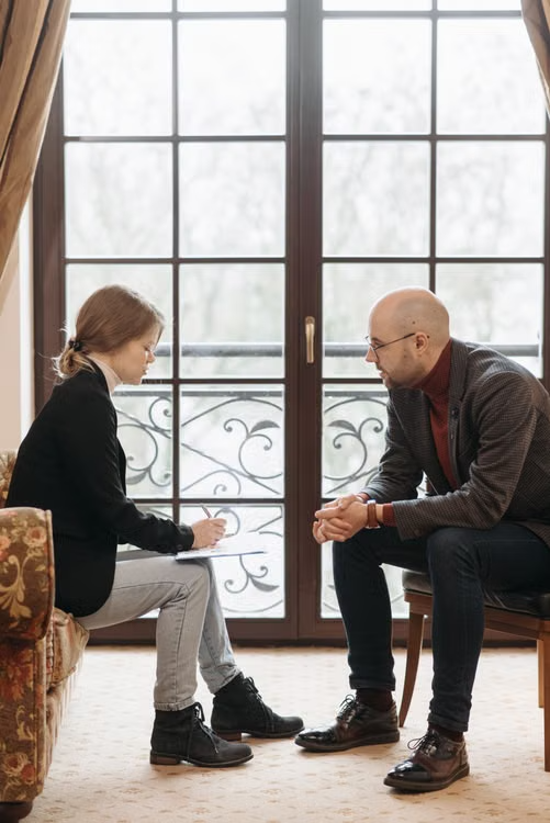 Two people seated facing each other in a quiet room by a tall window during a coaching conversation.
