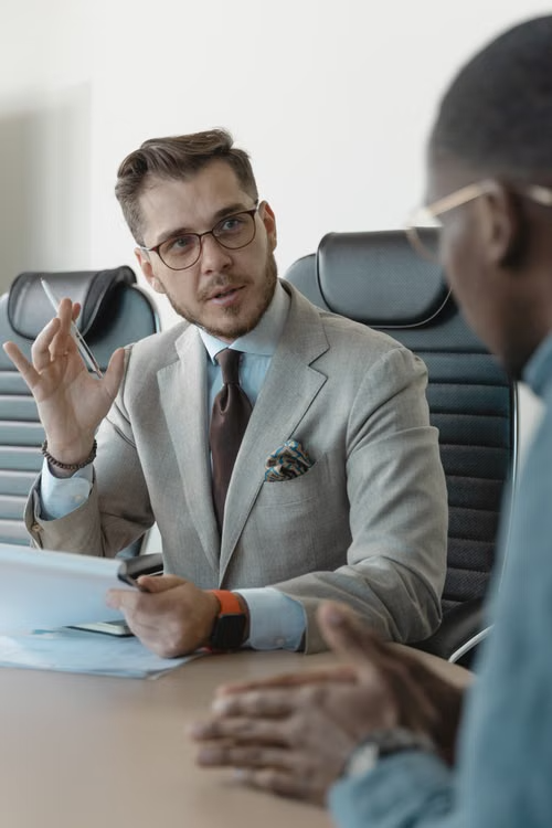 Two colleagues in conversation across a meeting table, one speaking and gesturing.
